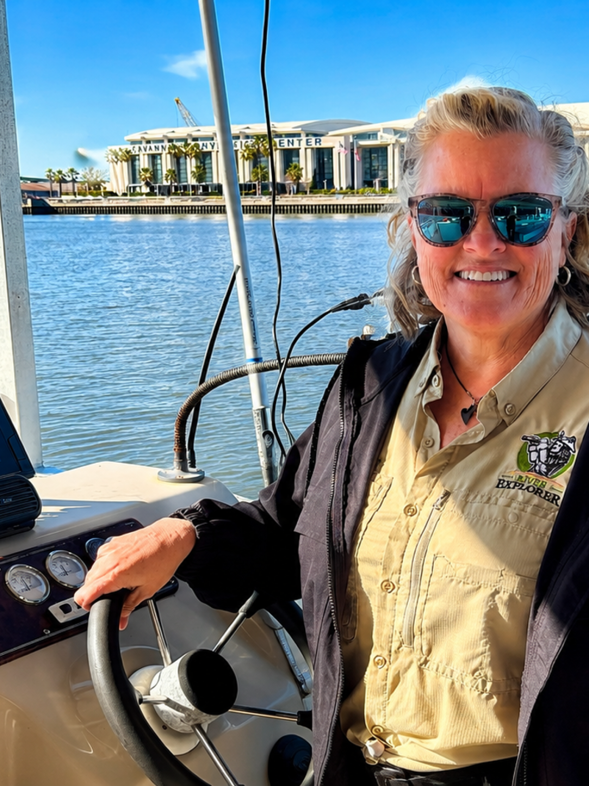 Smiling person in sunglasses steering a boat on a sunny day.