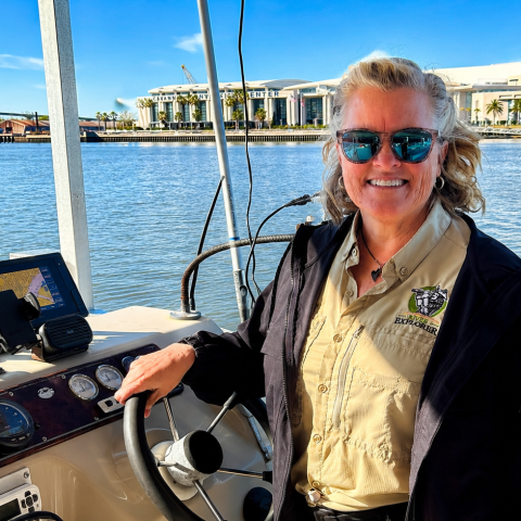 Smiling person in sunglasses steering a boat on a sunny day.
