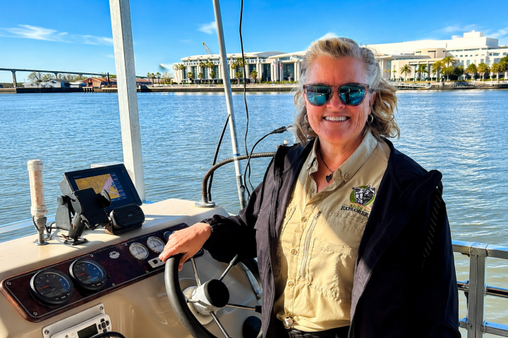Smiling person in sunglasses steering a boat on a sunny day.
