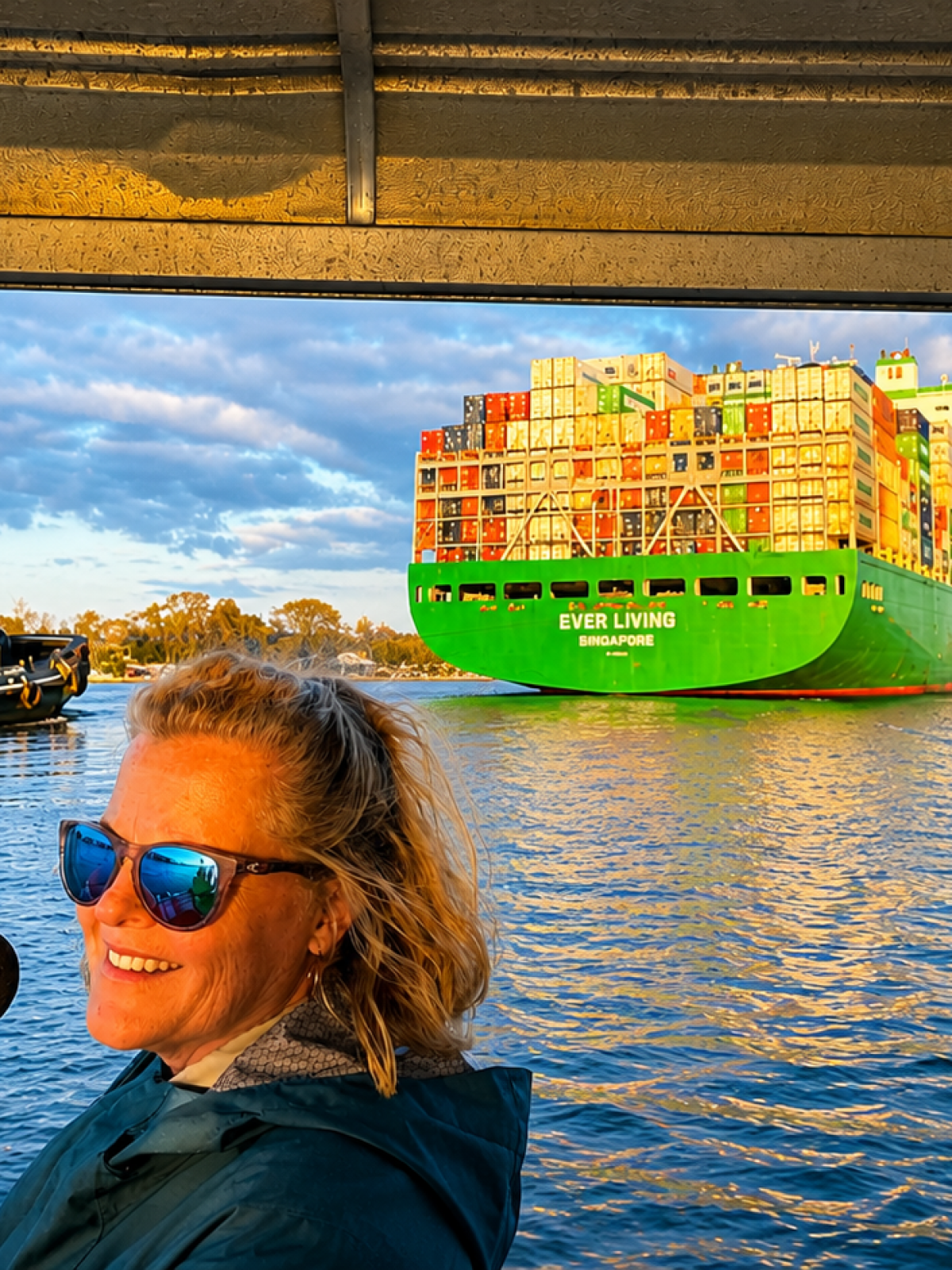 Woman at microphone on boat with cargo ship and tugboat on sunny day.