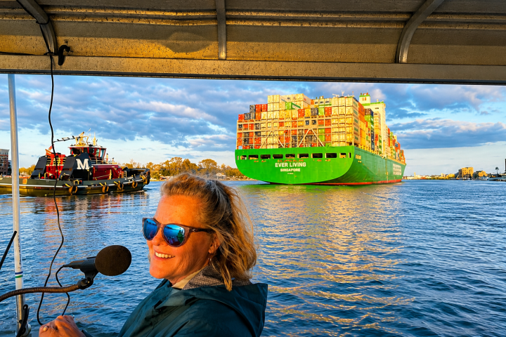 Woman at microphone on boat with cargo ship and tugboat on sunny day.