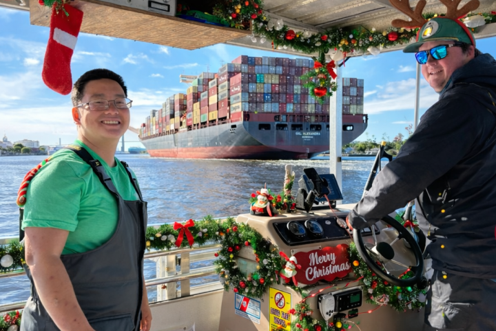 Two people on a decorated boat with a large cargo ship in background.