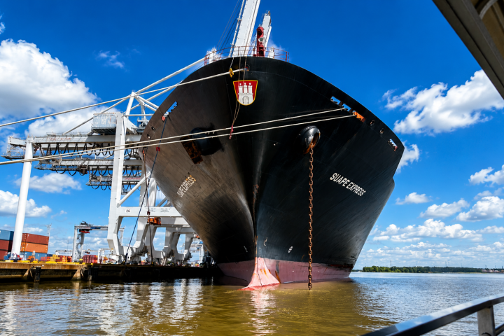 Large black cargo ship docked at port under a clear blue sky with white clouds.