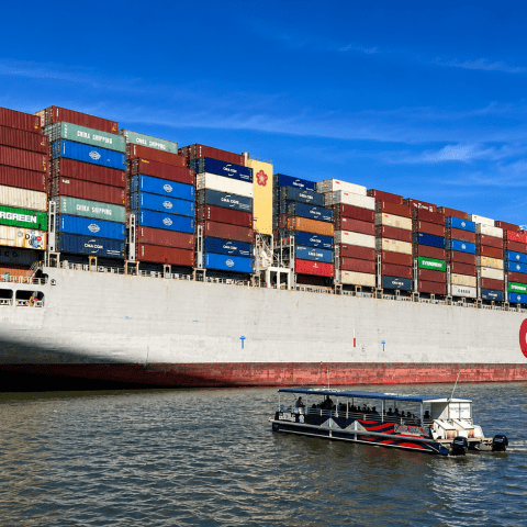 Large cargo ship with colorful containers, small boat in foreground on the water under a clear blue sky.