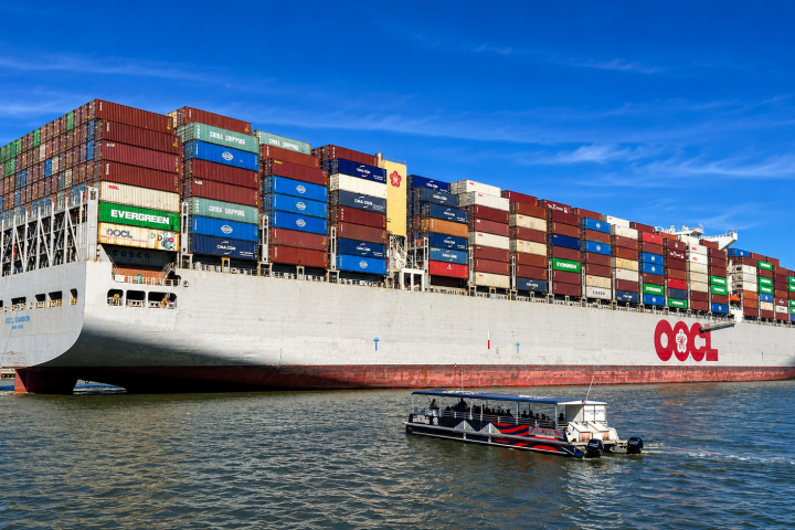 Large cargo ship with colorful containers, small boat in foreground on the water under a clear blue sky.