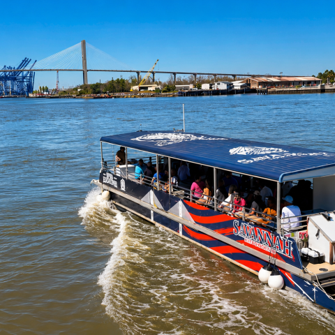 Boat on river with passengers, bridge and industrial area in background.