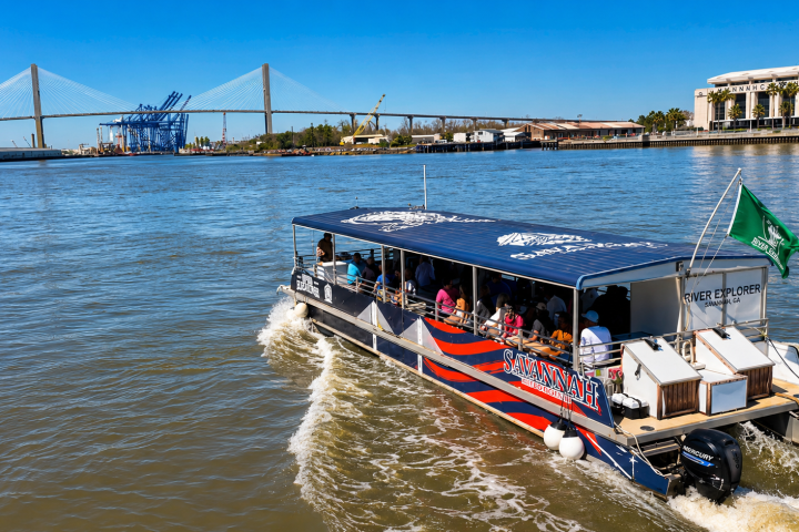 Boat on river with passengers, bridge and industrial area in background.