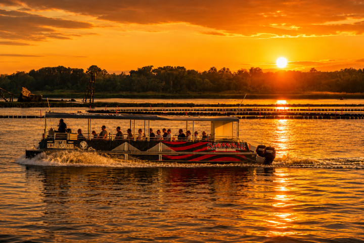 Tour boat on river at sunset with orange sky and reflections.