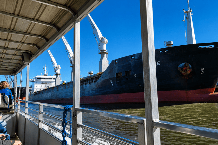 View from boat of large cargo ship with cranes on a sunny day.