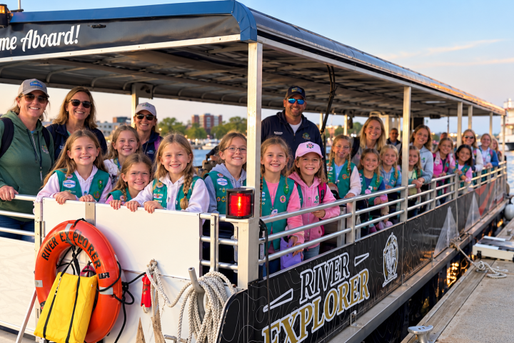 Group of uniformed girls and adults on a docked tour boat named 'River Explorer.'