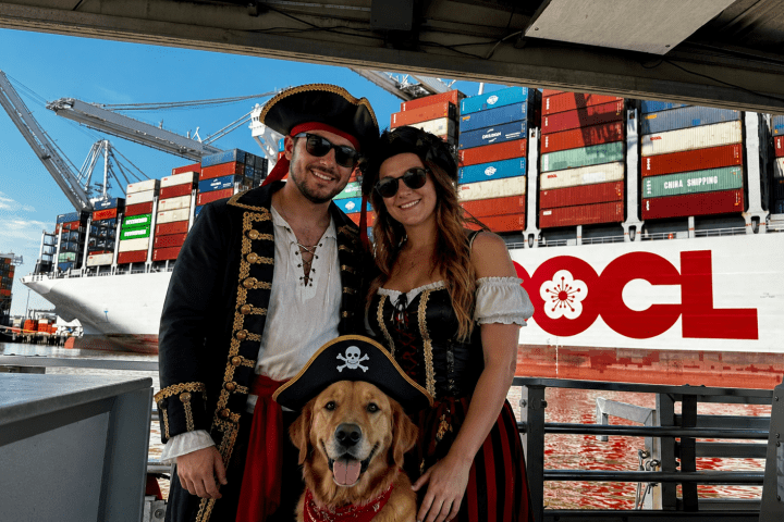Couple in pirate costumes with a dog, standing near a large container ship at a port.