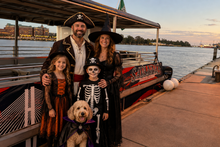 Family in pirate costumes with dog on riverside dock at sunset.